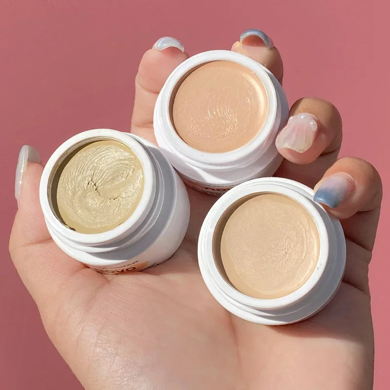 Three open white containers of cream makeup held by a hand against a pink background